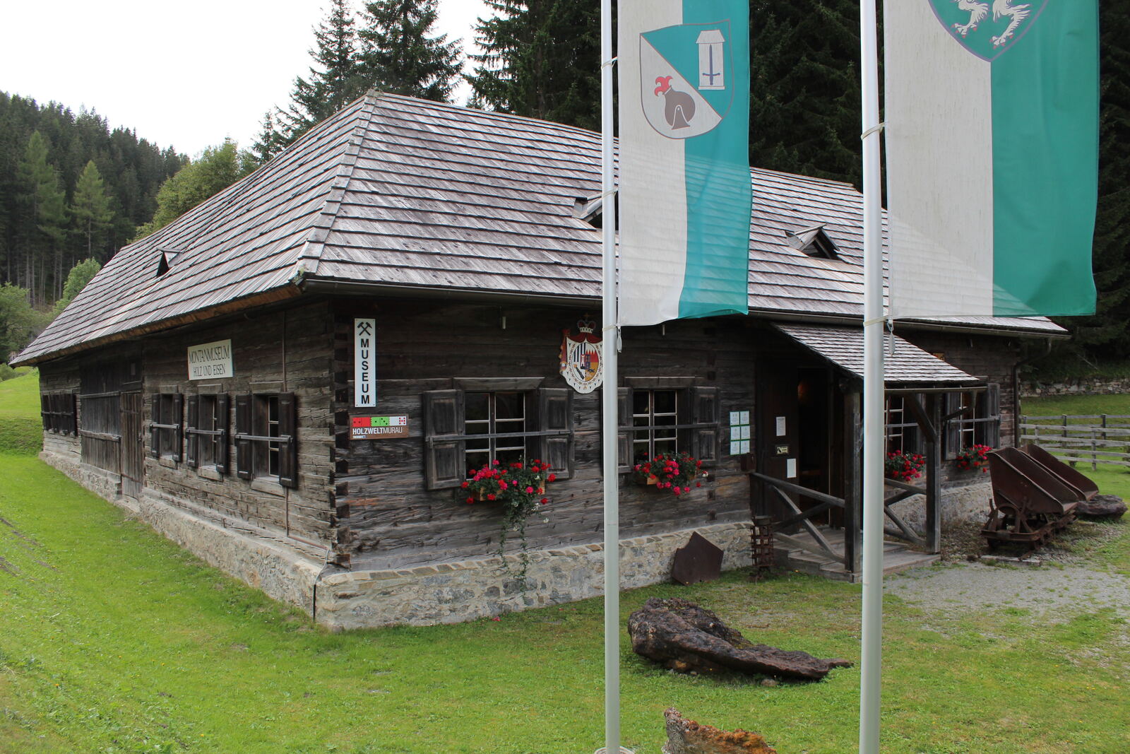 Das Montanmuseum Holz und Eisen – altes Blockhaus mit Schindeldach, davor Hunte und Fahnen